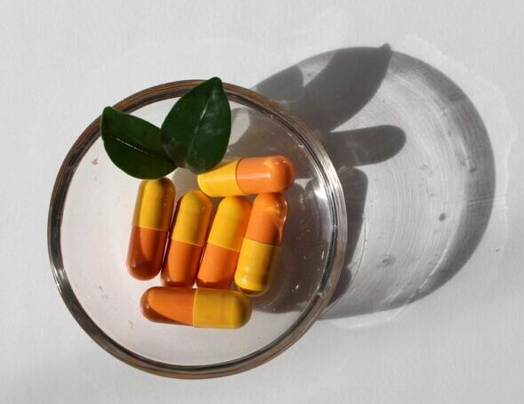 capsulas de remédio amarelas e laranjas dentro de um copo de vidro e folhas verdes