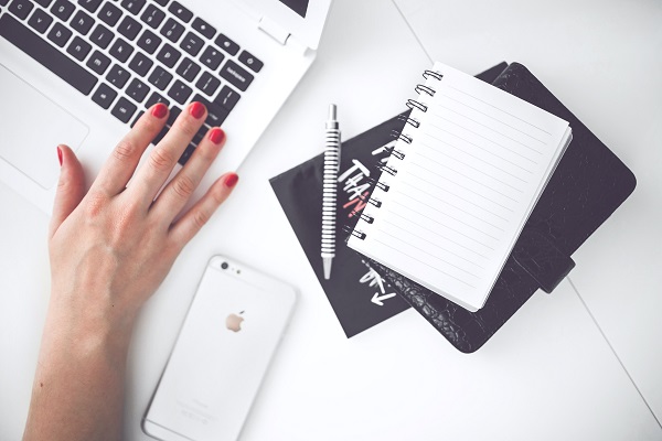 uma mão digitando no computado com unhas vermelhas e ao lado caderno e caneta