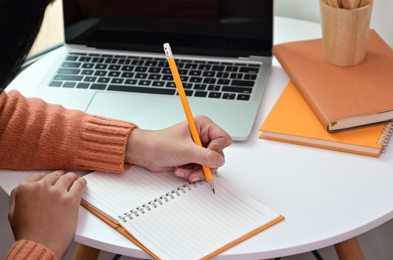 Mulher com blusa laranja escrevendo em um caderno com um lápis amarelo